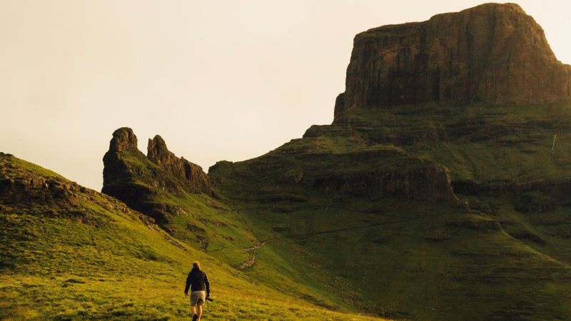 Drakensberg walker crosses grassland beneath a rugged peak, South Africa - Drakensberg Walking Tour, South Africa